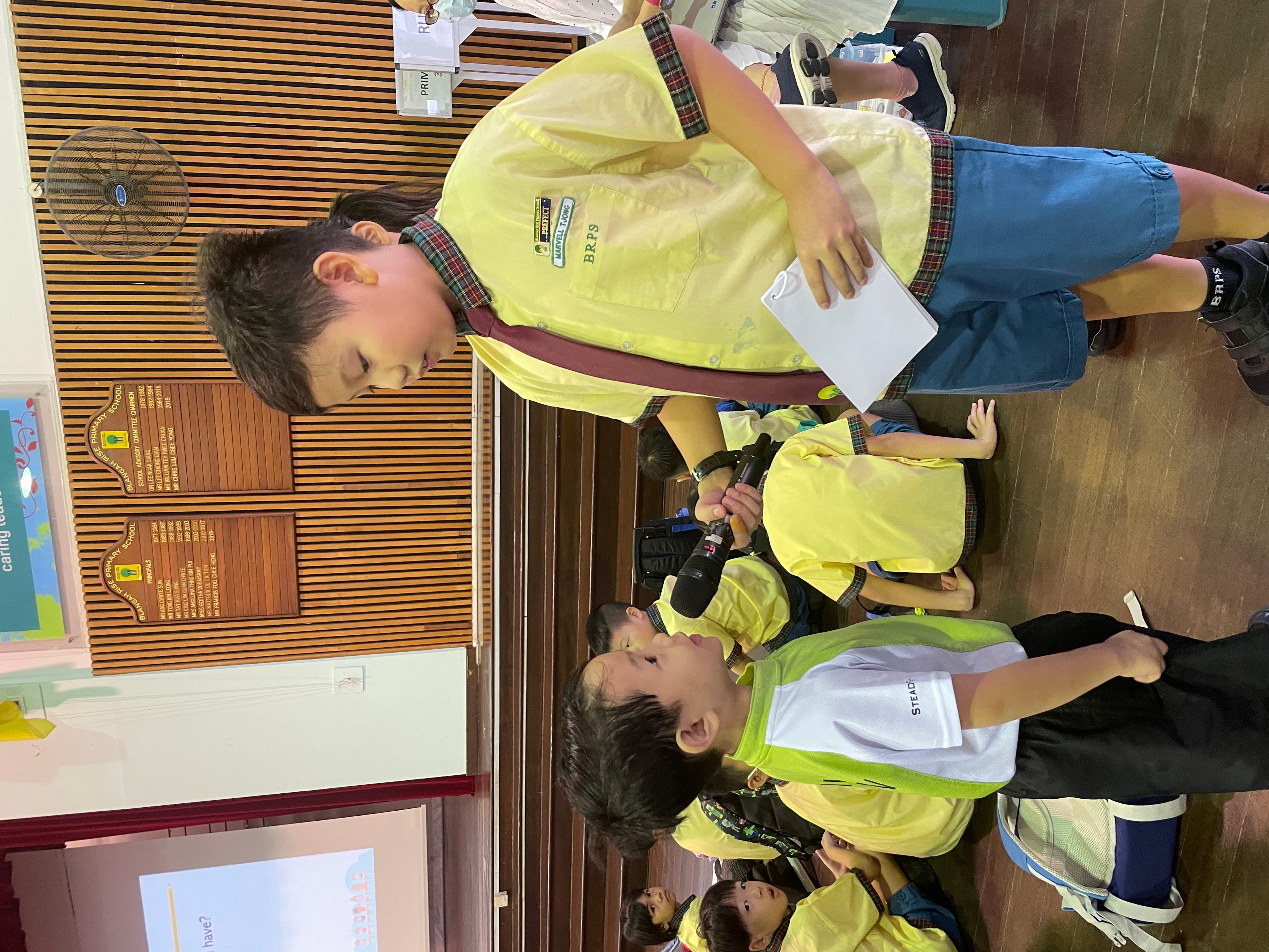Boy holds a microphone to another boy wearing a green vest, while other kids sit on the floor behind.
